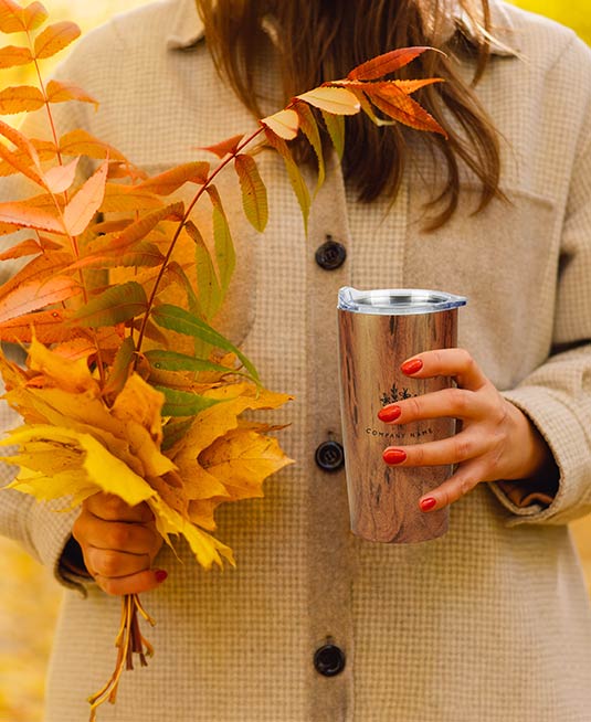 Person holding a wood tone tumbler that displays a logo in one hand, and a bouquet of fall leaves in the other hand.