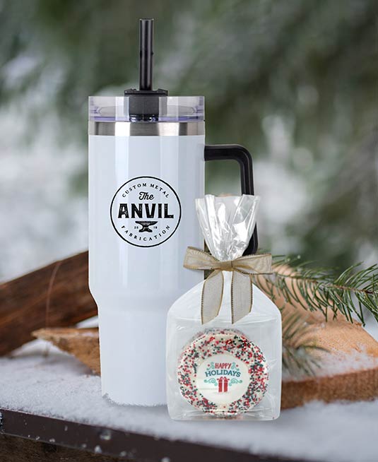 Stanley style tumbler with a custom logo sitting on a table outside in the snow with an individually wrapped custom cookie.