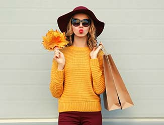 Person in a yellow shirt, sunglasses and a hat, holding a shopping bag in one hand and orange leaves in the other.